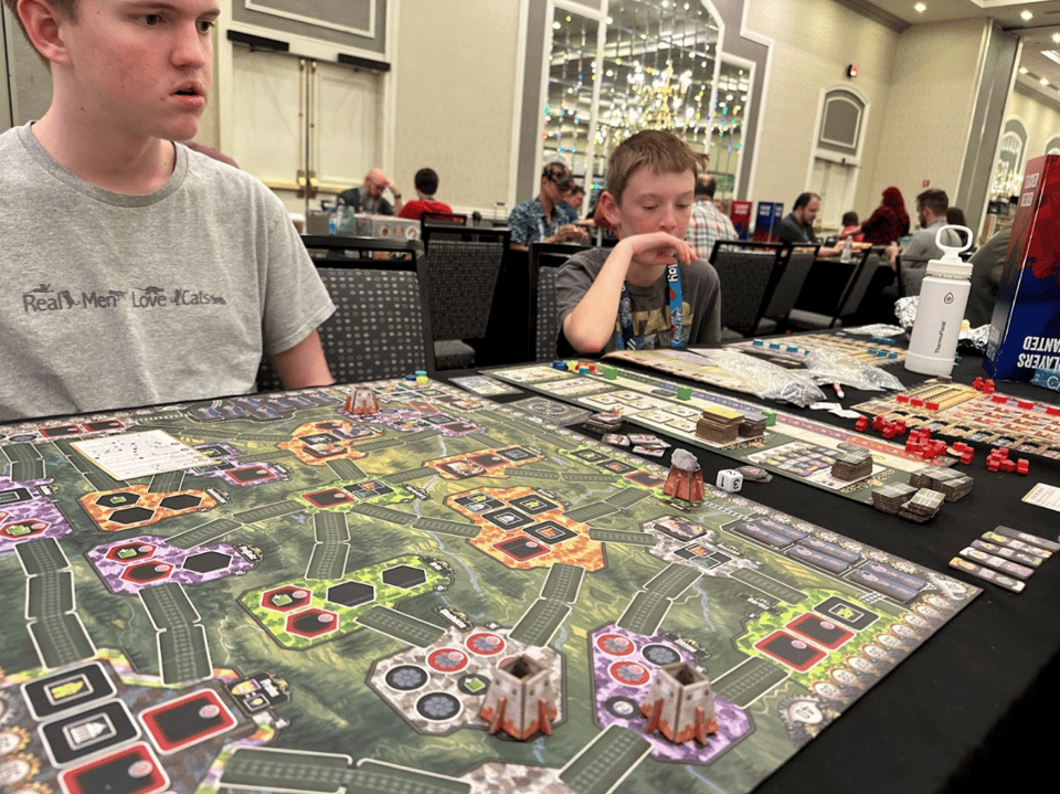 Players look at a game board at a long table in a convention hall