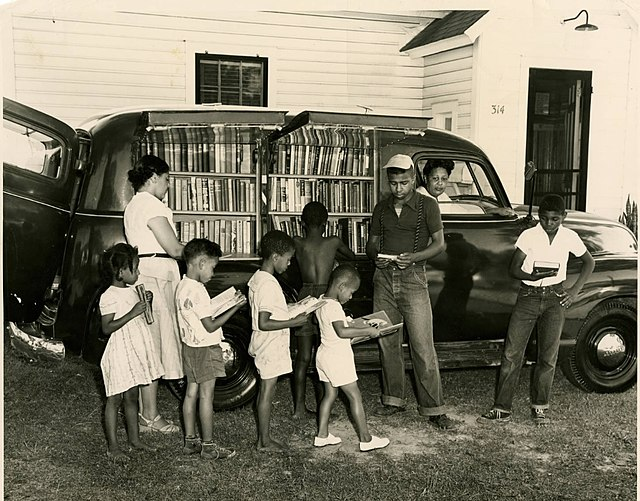 African-American children secure books at a North Carolina Albemarle Region bookmobile stop. Black woman is in the front seat of the bookmobile.