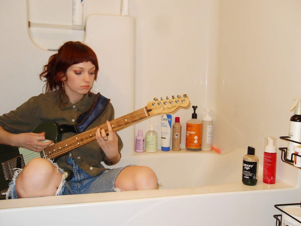 A young woman with dyed auburn hair wearing a dark green buttondown shirt with the sleeves rolled up and jeans with ripped knees crouches in a bathtub while playing a black Fender Telecaster with a white pickguard.