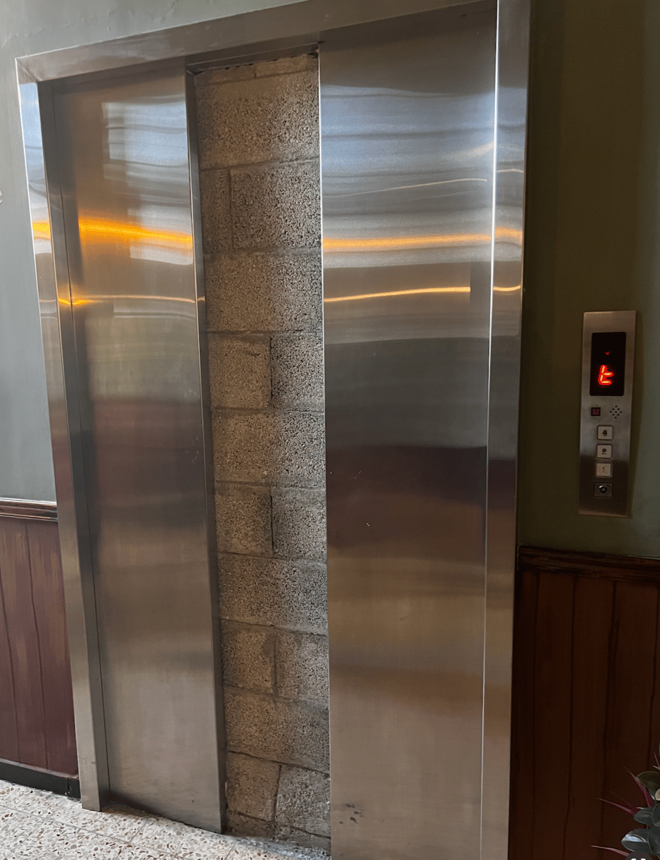 A photograph of an elevator next to a wall with a half-height wood panel. The stainless steel doors of the elevator are slightly open, exposing a gray cinderblock wall right behind the doors. The elevator control panel to the right displays what looks like an 'E' with a missing top line; or perhaps it's a lowercase 't'.