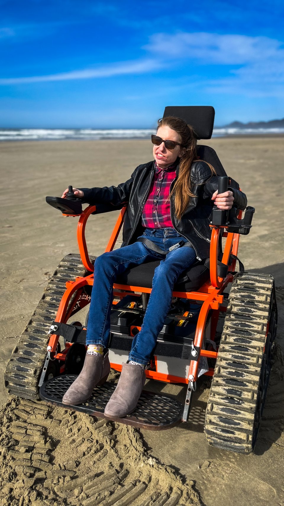 A woman with reddish-brown hair, dressed in a leather jacket and flannel shirt, sits in a bright orange all-terrain wheelchair on a sandy beach. She faces toward the sun on a clear day.