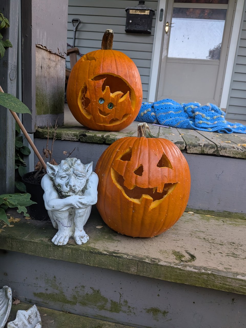 The front porch of a house. On the steps are two jack-o-lanterns next to a small stone gargoyle whose wings have broken off.