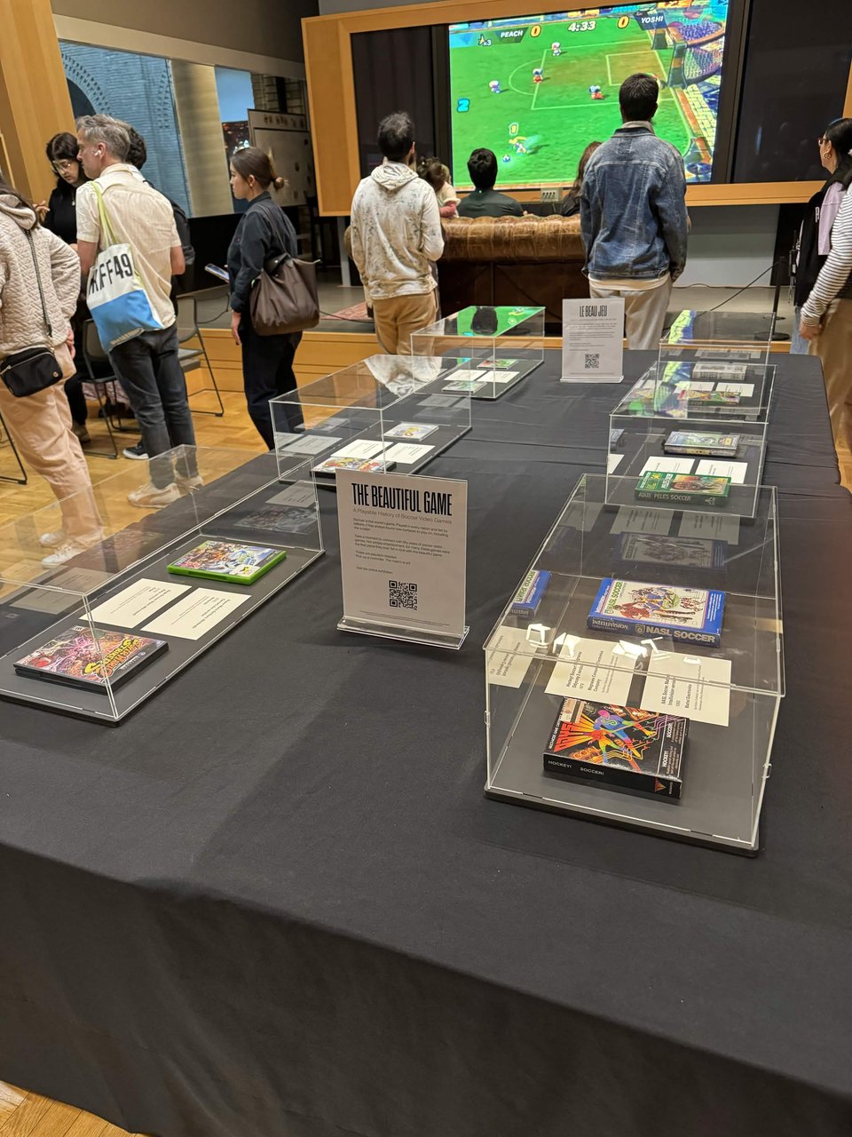 A museum exhibit titled "The Beautiful Game: A Playable History of Soccer Video Games," where vintage soccer video game boxes are displayed under clear acrylic cases on a long table draped in dark grey cloth, while in the background visitors stand watching a large screen showing a Super Mario soccer video game.