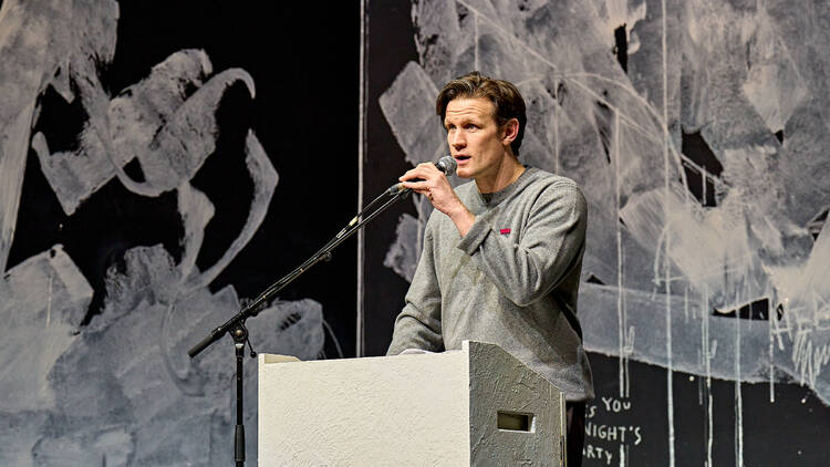 An image of Matt Smith in An Enemy of the People at Duke of York's Theatre. He is standing at a white podium, holding and talking into a microphone attached to a stand. He is wearing a grey jersey. He is standing in front of a black wall with lots of white paint splattered over it.
