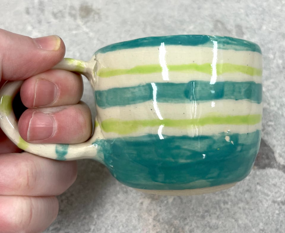 A tiny teacup, hand-thrown on a wheel, painted with chartreuse and blue stripes