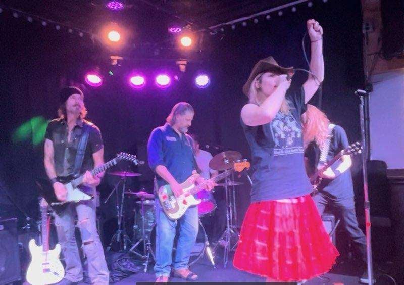 A woman in a red skirt, t-shirt and cowboy hat sings into a microphone in front of a four-man backing band comprising (from left) guitar, bass, drums and guitar.