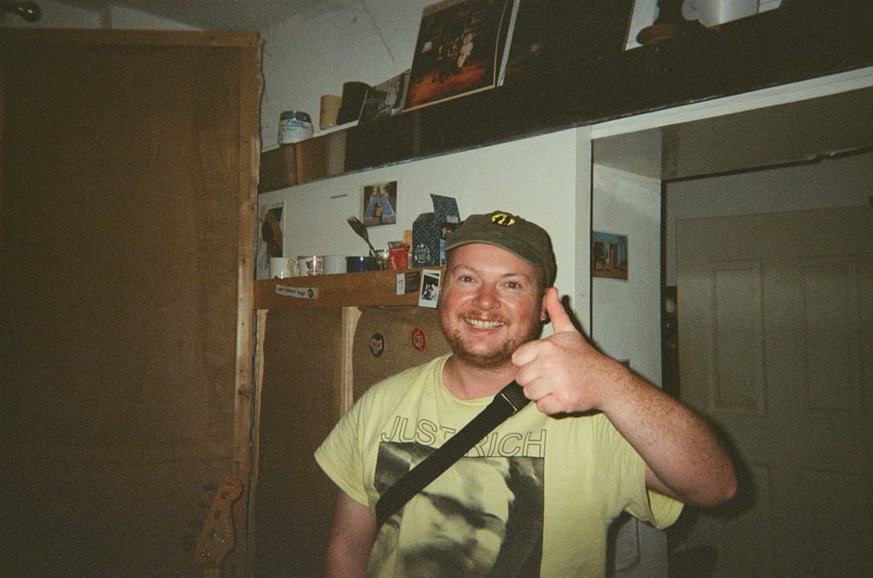 Man smiling and giving a thumbs up. He is wearing a cap and a yellow t shirt