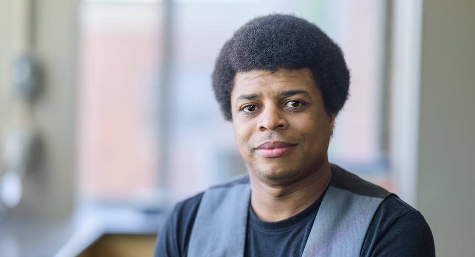Photo shows JMC Assistant Professor Orlando Hawkins, photographer indoors wearing a back shirt and grey vest.