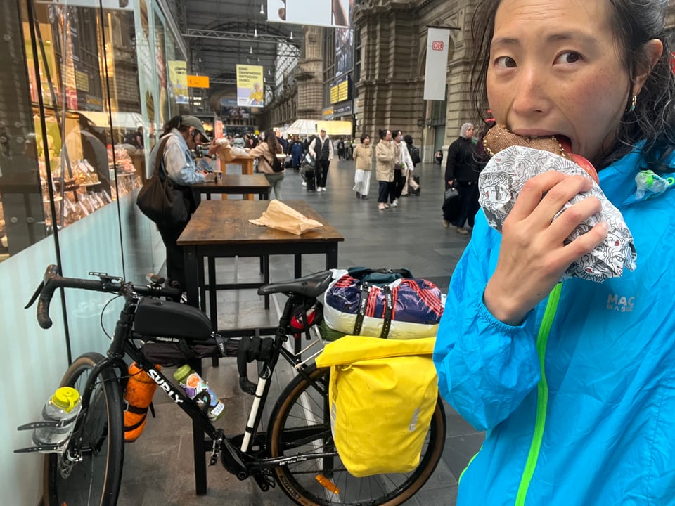 Jenny looking tired and wet, eating a sandwich, with laden bike and busy train terminal in background.