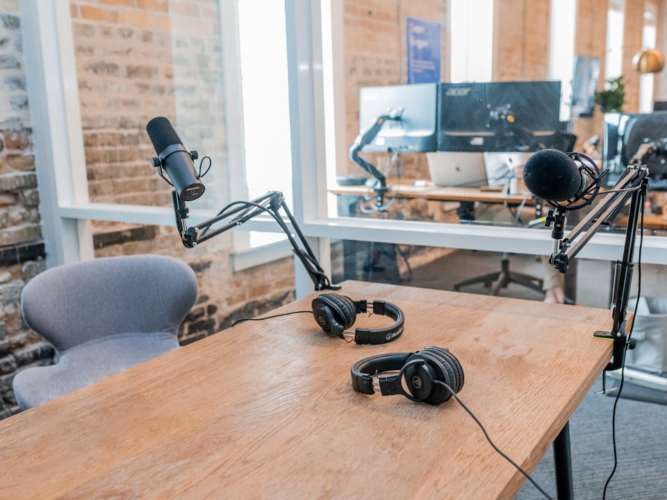 Photo of what appears to be an in-office podcast studio: a wooden table, with a microphone and set of headphones on either side. An office environment is visible in the background, through a glass wall. Photo by Austin Distel on Unsplash.