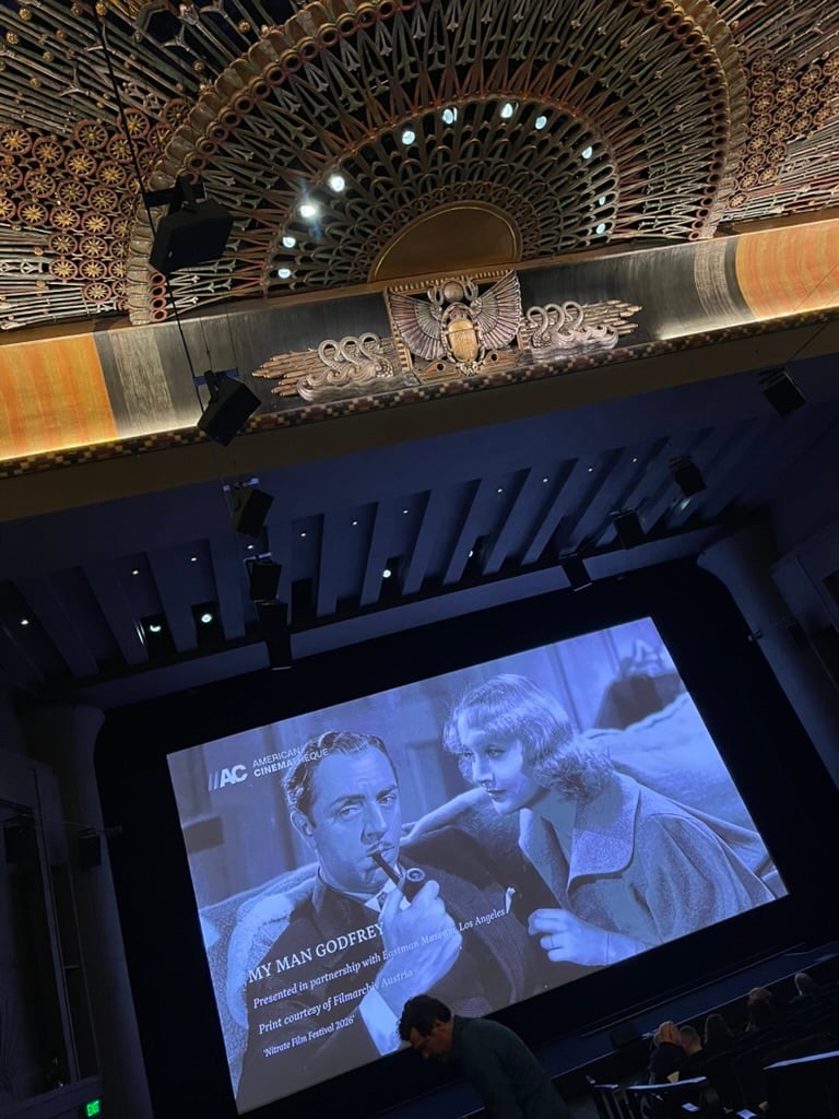 An ornate theater. On the screen is a big ad for the film My Man Godfrey. People mill about finding their seats.