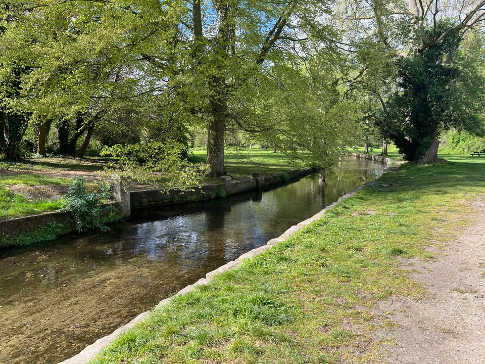 A shallow river running through a green park. The river is quite wide with stone walls on either side, grass on the banks. The bottom of the river is clearly visible.