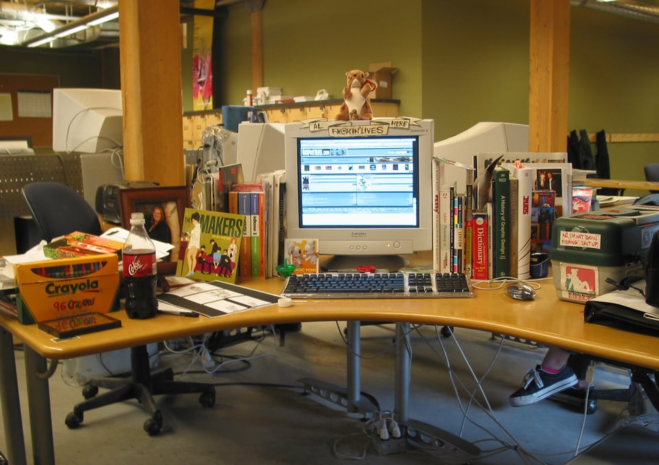 A picture of Allan Lorde's third-year graphic design program workstation at Red River College in Winnipeg, Manitoba. At his desk are assorted books, a tackle box full of supplies, a box of Crayola crayons, a bottle Cherry Coke, and a 2000s era Mac computer. Taken September 15th, 2004.