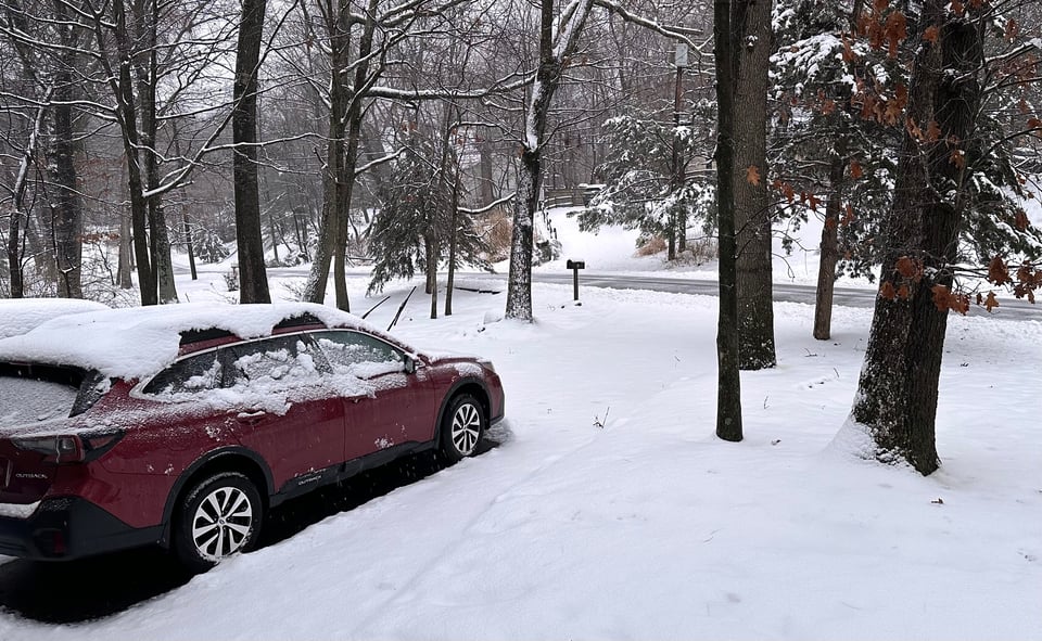 Photo of front yard and driveway w/dark-red Subaru Outback, all covered by snow