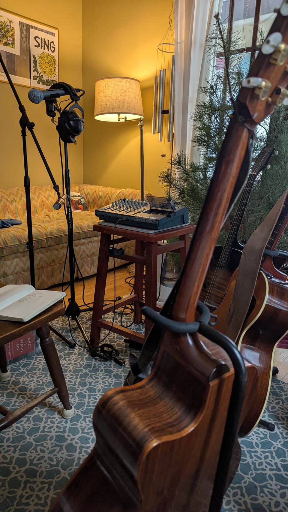 My practice studio (a.k.a. the parlor), set up like an open-mic stage wanted to cut a record, with a chair (empty save for a notebook, open to a set list), two dynamic mics (one high, one low) plugged into an ancient TEAC Porta-Studio recorder, and three kinds of guitars off to one side. Comfy furniture and the stubby remains of my holiday tree are in the background.