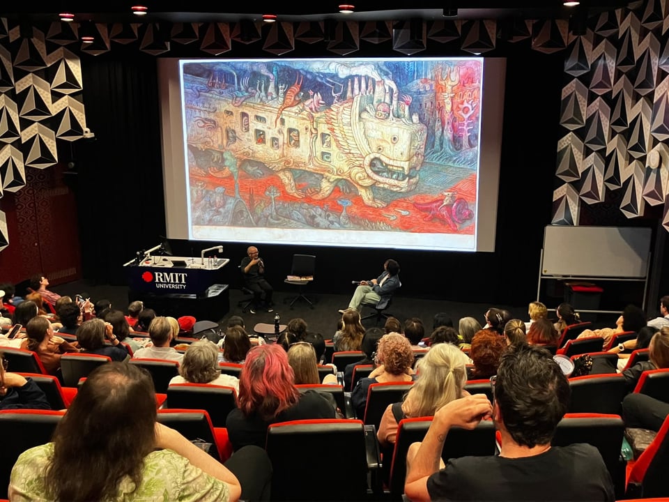 An audience seated in a dark RMIT University lecture theatre watches Shaun Tan and Campbell Whyte on stage beside a large projection screen displaying colourful, surreal art from Shaun Tan's Hometown graphic novel.