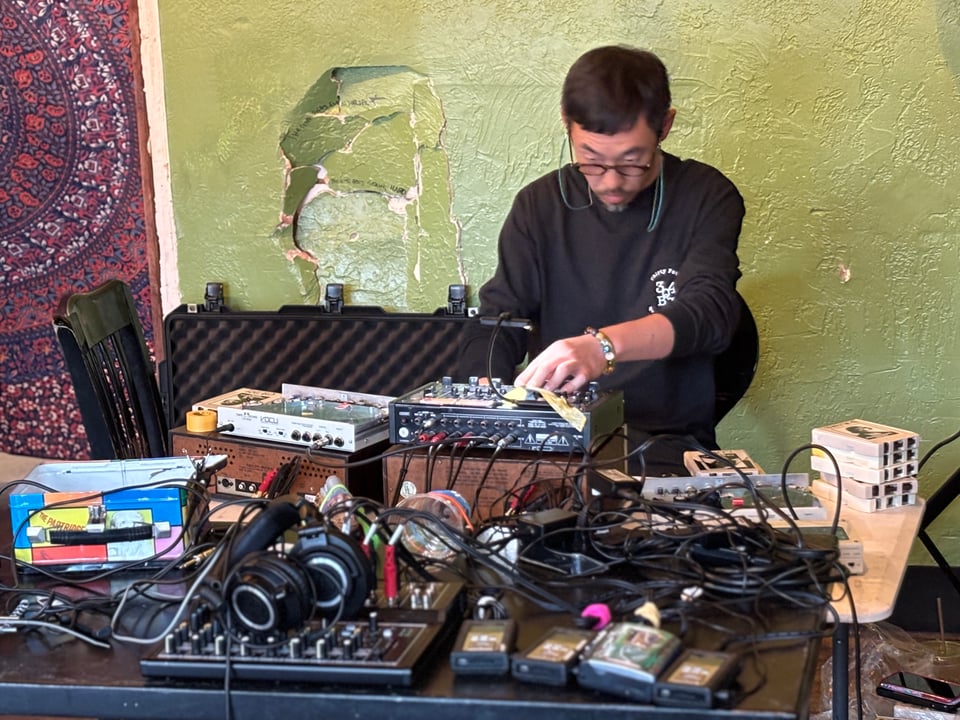 Shin Chida performing live, sitting at table covered in tape machines and effects pedals