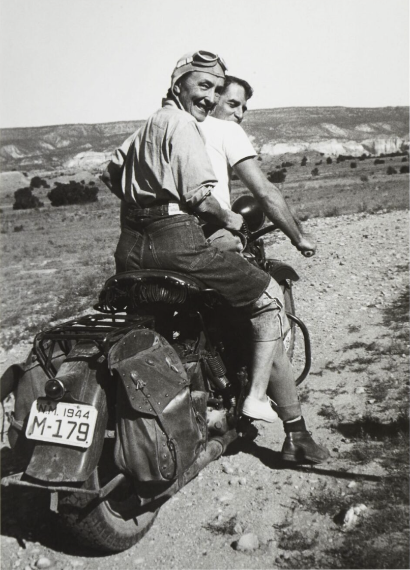 Georgia O'Keeffe smiling on the back of a motorcycle being driven by a younger man