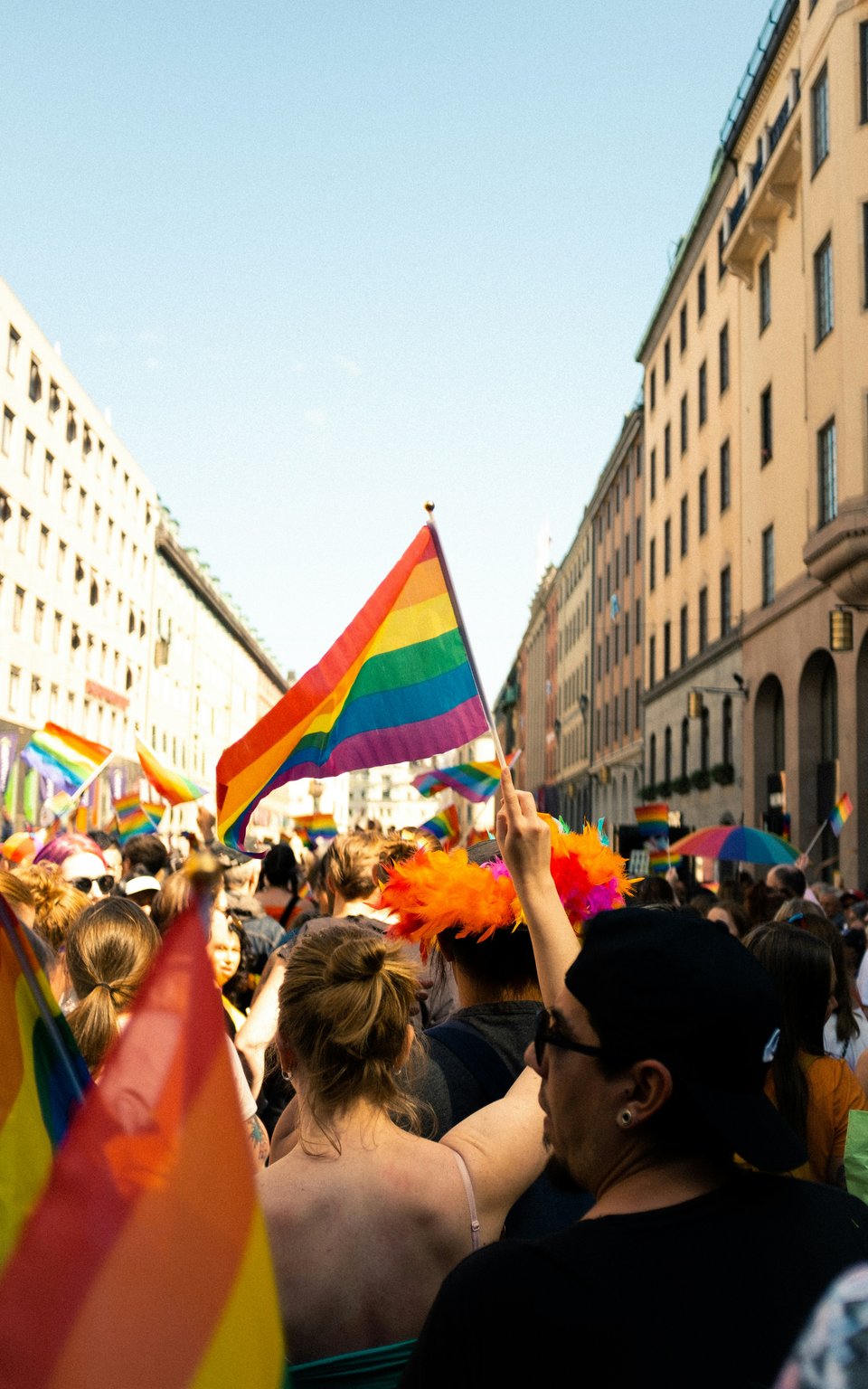 A crowd celebrating Pride outdoors. Many people waving flags outside on a city street on a sunny day.