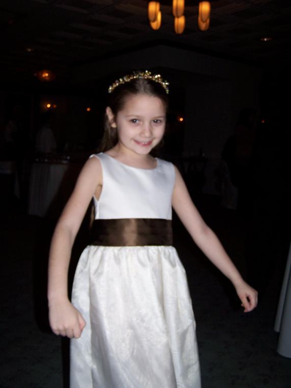 Photo of a 6-year-old white girl in a white gown with a brown belt and a tiara, at her uncle's wedding
