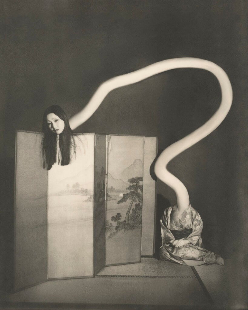 A sepia-toned photograph depicting the Japanese yokai "rokurokubi": a kneeling woman in a patterned kimono sits beside a folding landscape screen, her neck stretching upward in a long, sinuous white curve that arcs over the screen to a pale, dark-haired head floating above its left panel.