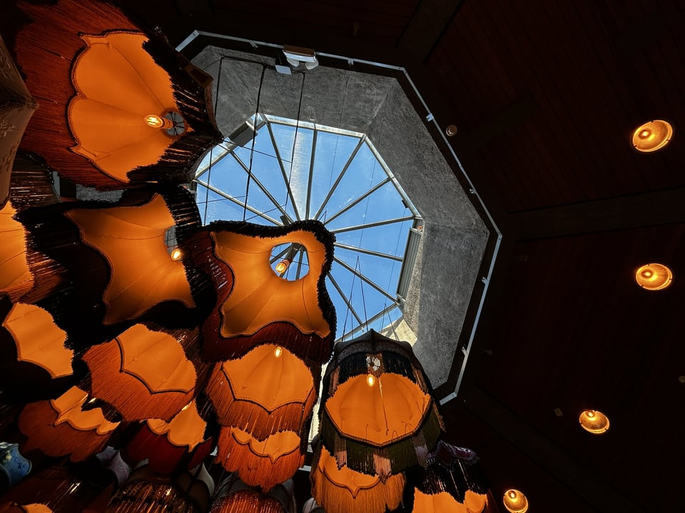 A concrete octagonal lantern set within a wood paneled ceiling and half-hidden by a large set of orange 1970s fringed lampshades clustered together.