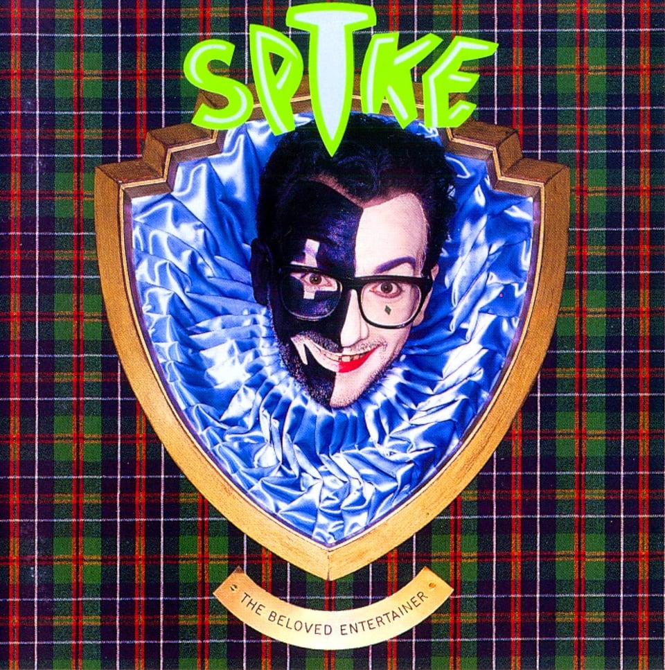 Elvis Costello, with his trademark thick-rimmed black glasses, his head poking through a shield-shaped plaque. Within the shield, his head is surrouned by blue ruffles of some kind of fabric. Costello looks like a harlequin; half his face is painted black, the other half white. The black side has a white strip horizontally over his eye, and his lips are painted white as well. On the white side, his lips are painted red. He smiles maniacally. The plaque has a plaid background. The plaque is labeled "THE BELOVED ENTERTAINER."