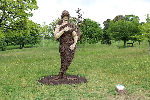 A willow or wicker sculpture of a woman with a seal's body stands in a green field.