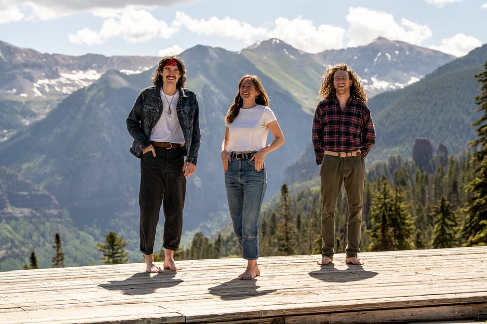 A barefoot woman in a white t-shirt and jeans stands between two men with long hair, also barefoot, on a wooden platform in from of mountains.