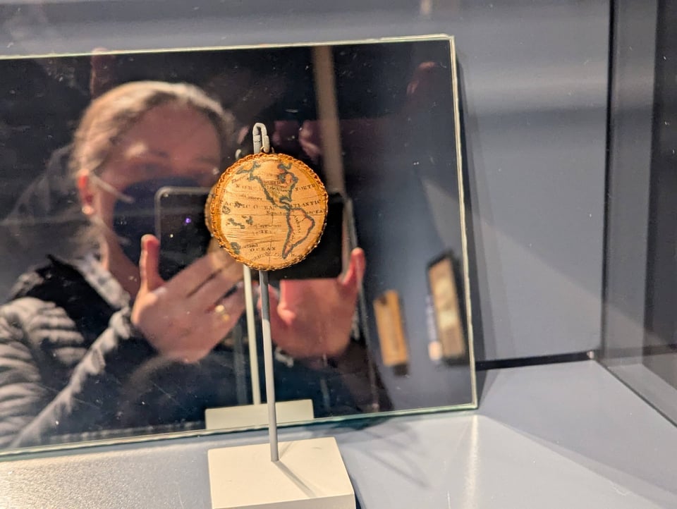 My masked face and camera appear in a mirror behind an embroidered circle showing a nineteenth-century map of the Americas