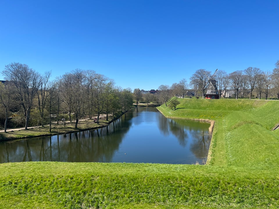 Looking down into a moat from atop a grassy island. The slopes of the island are made entirely of straight lines and form some sort of angular shape. On the far bank are some trees and a footpath.