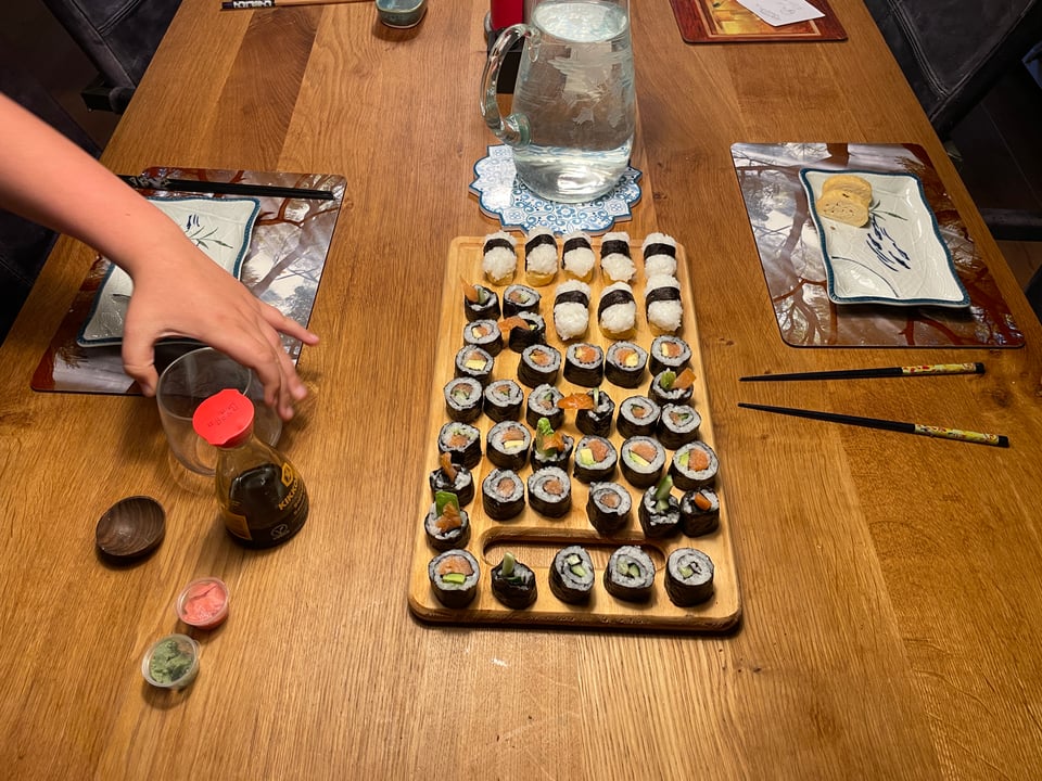 A table is set with two plates, chopsticks, glasses and a cutting board filled with homemade sushi.