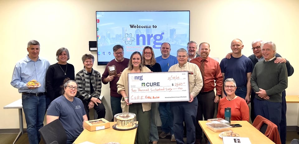 A group photo of members and guests in front of the NRG welcome screen. Bruce and Holly hold a big check made out to CURE for $2,660.