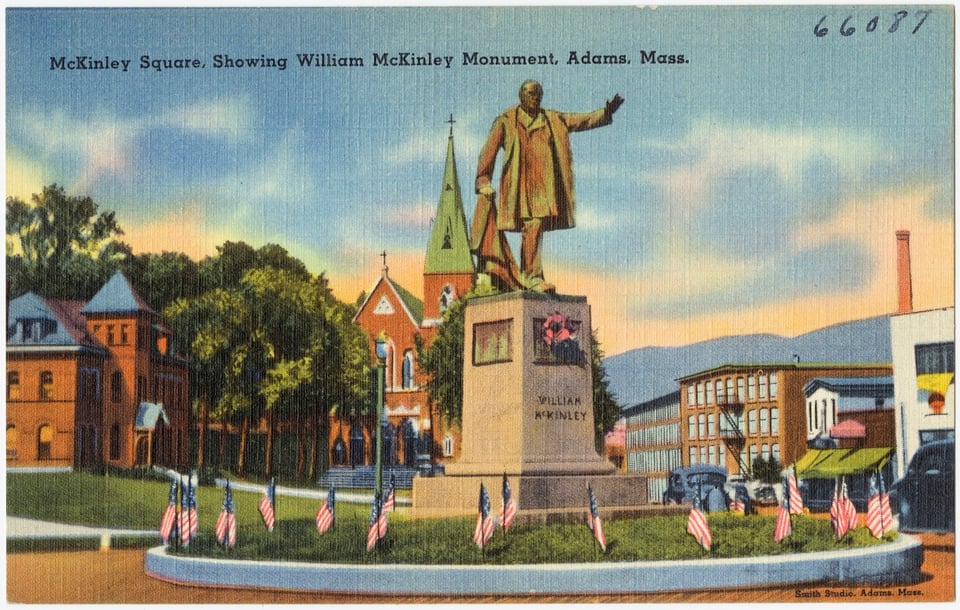An old-timey postcard from the 1930s offers a sepia view of downtown Adams, with the McKinley state in front surrounded by little flags