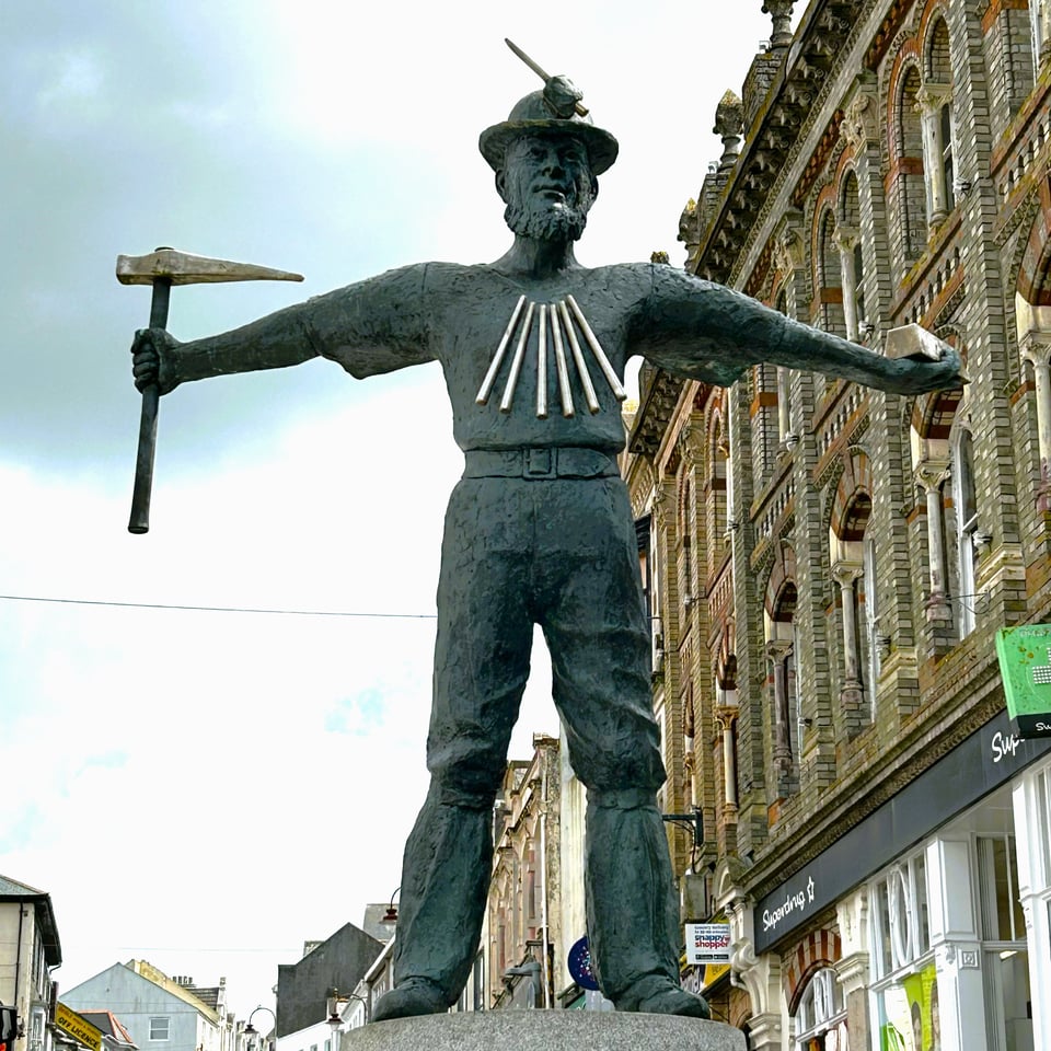 A 2m high bronze sculpture of a tin miner. He's standing like Leonardo De Vinci's Vitruvian Man, but clothed. In one hand he holds a mining adze and in the other an ingot of tin. He has metal candles around his neck, and a candle stump on his hard hat.
