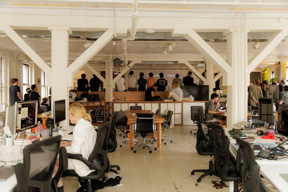 In the foreground are two rows of desks with a couple people working. In the background people watch an event, the projector screen just visible.