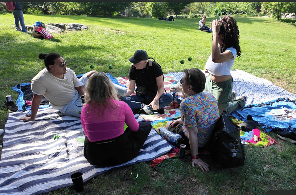 John Biewen seated on blankets on a grassy lawn, holding a microphone, listening to a diverse group of four younger people seated around him.