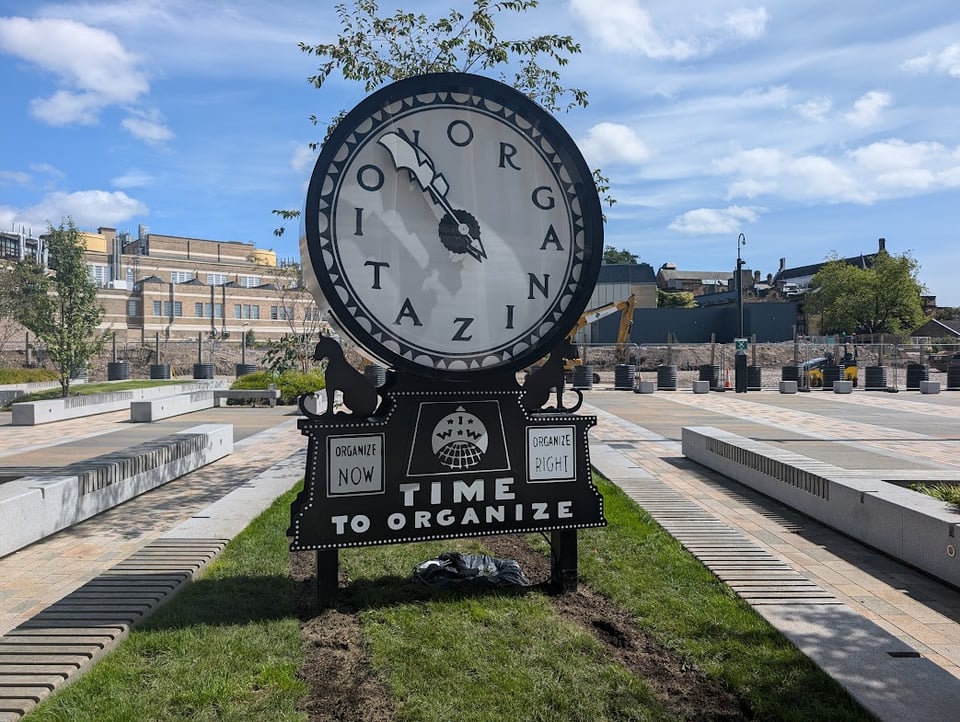A big black and white clock, except that instead of numbers it has the letters that spell "ORGANIZE" Underneath is a union logo and hte words "Organize Now" "Organize RIght" "TIME TO ORGANIZE". There are cute animals on the sides. It's on a college campus in some kind of quad.