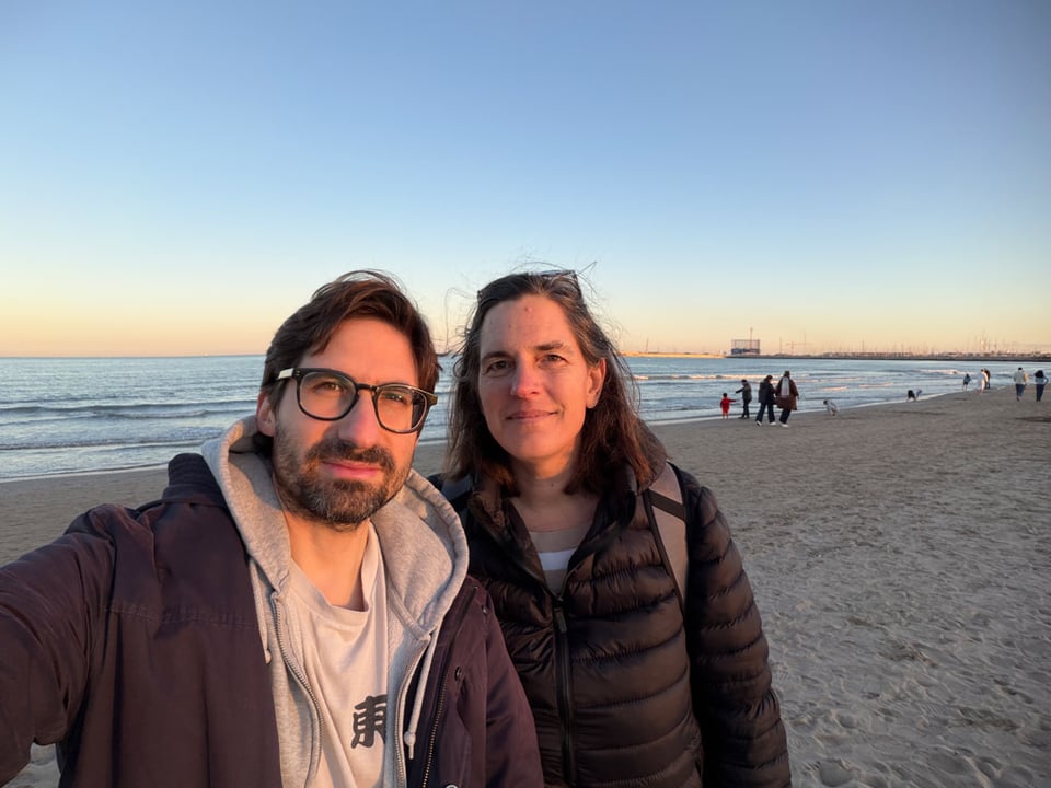 Two people look at the camera wearing jackets on the beach with the ocean behind them.