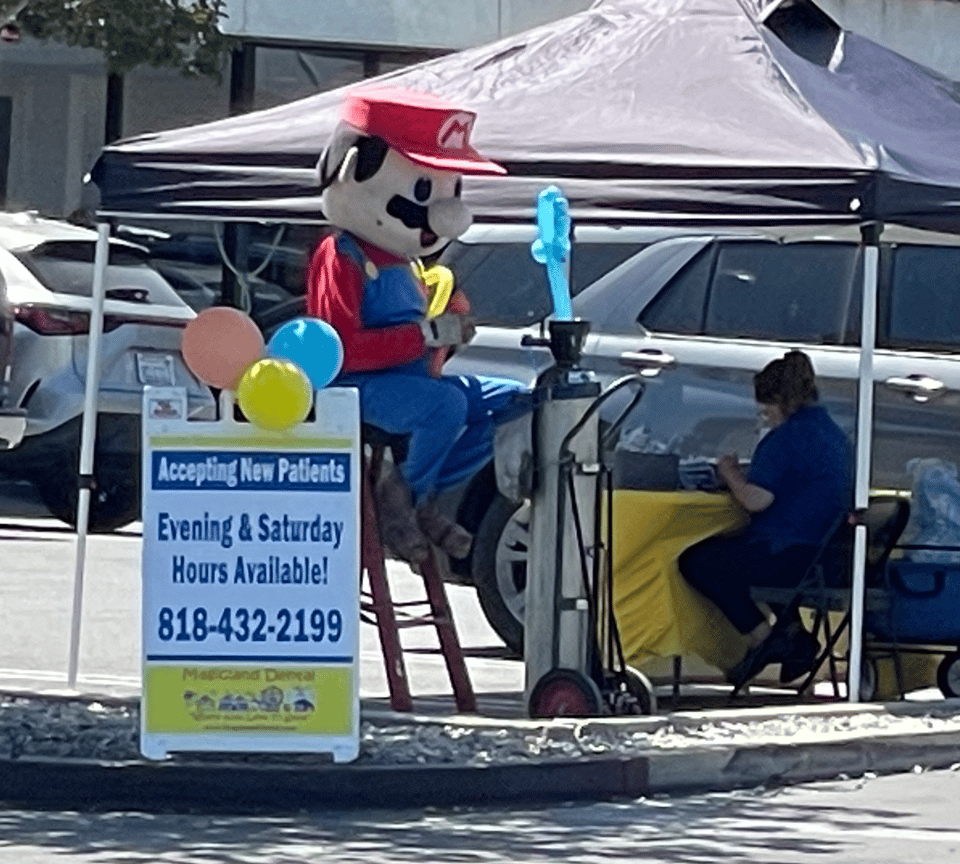 A person dressed as Mario sits by a DJ and a balloon machine in a parking lot