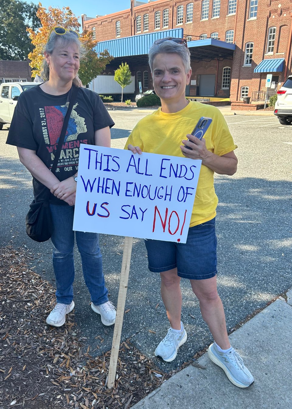 Photo of two people on sidewalk holding sign that says: "THIS ALL ENDS WHEN ENOUGH OF US SAY NO!"