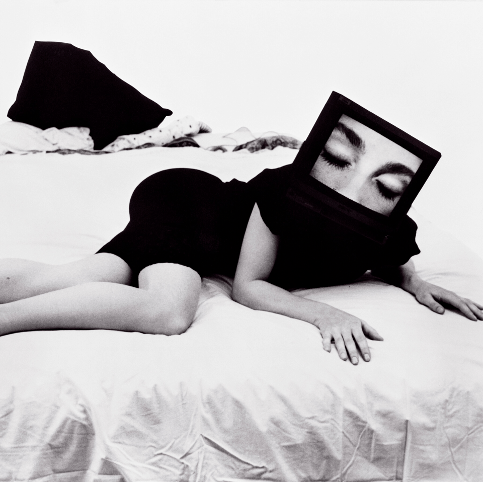 a black and white photo of a woman reclining on a bed whose head has been replaced by a large television set displaying an image of a woman's face