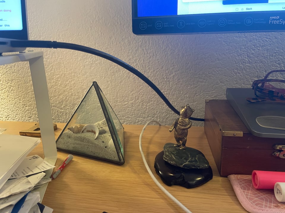 A desk covered in knick knacks. There is a pyramid of glass with sand and seashells inside. Next is a bronze statue of winnie the pooh on a piece of jade stone.