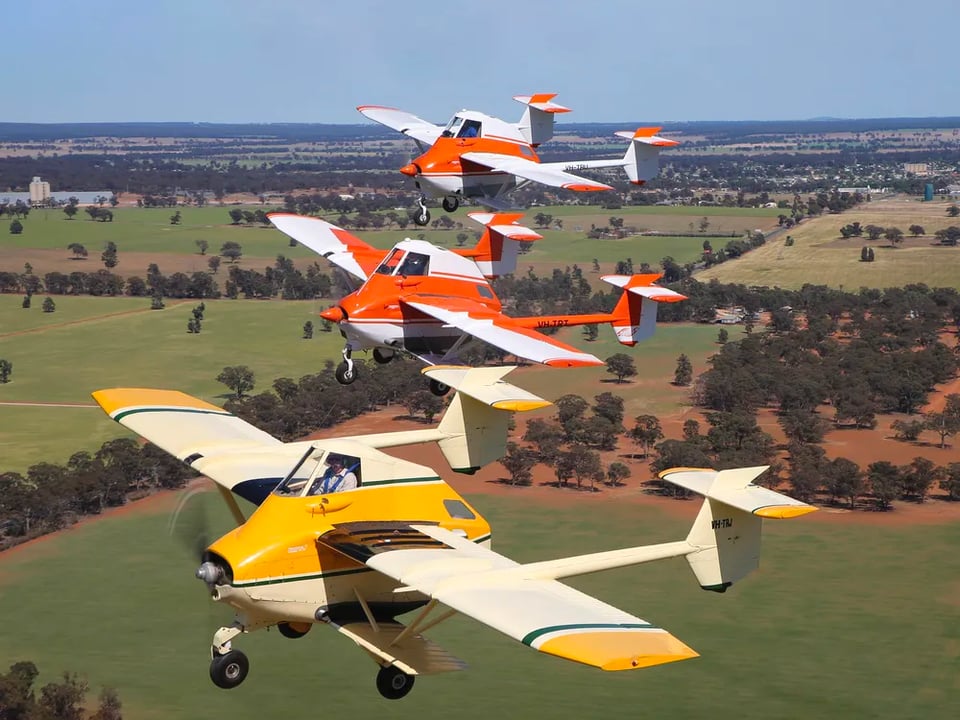 Three AirTruks flying in formation. Each plane has a tall, wide, stubby body, with the pilot seated up high and to the front. It has a propellor in the front. Each has two long winds sticking out from the sides of the body, as well as two smaller biplane-style shorter wings below. Each plane has one landing wheel up front and two behind, non-retractible. Each has two long arms jutting backwards from each wing, each with stabilizers. They are flying low above what appears to be farmland with lines of trees. The photo is taken from an angle to the side and slightly above the three planes