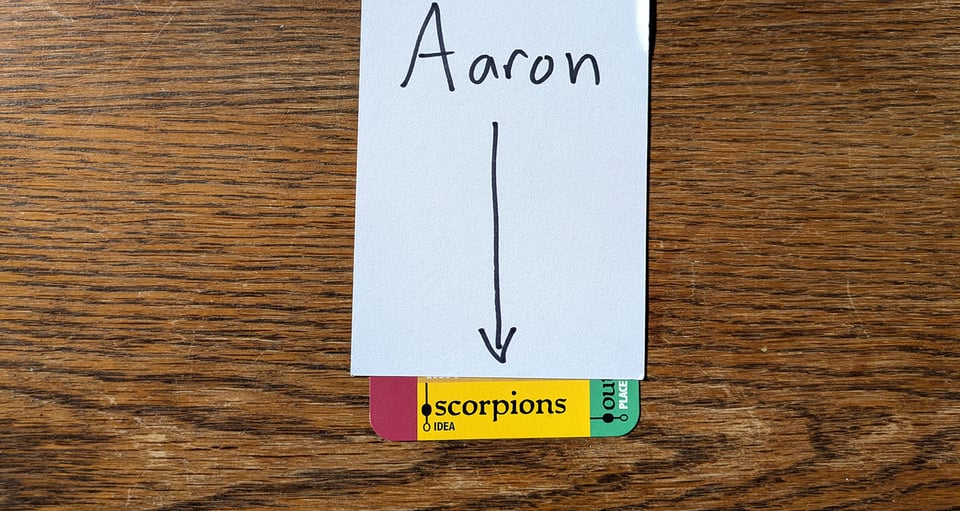An index card with "Aaron" written on it and an arrow, covering and pointing to the single word "scorpions" on a Downcrawl Deck card.