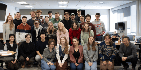 A group of students and a JMC professor are posing for a photo inside a Case Hall classroom. They are arranged in multiple rows, with some standing, others kneeling, all facing the camera.