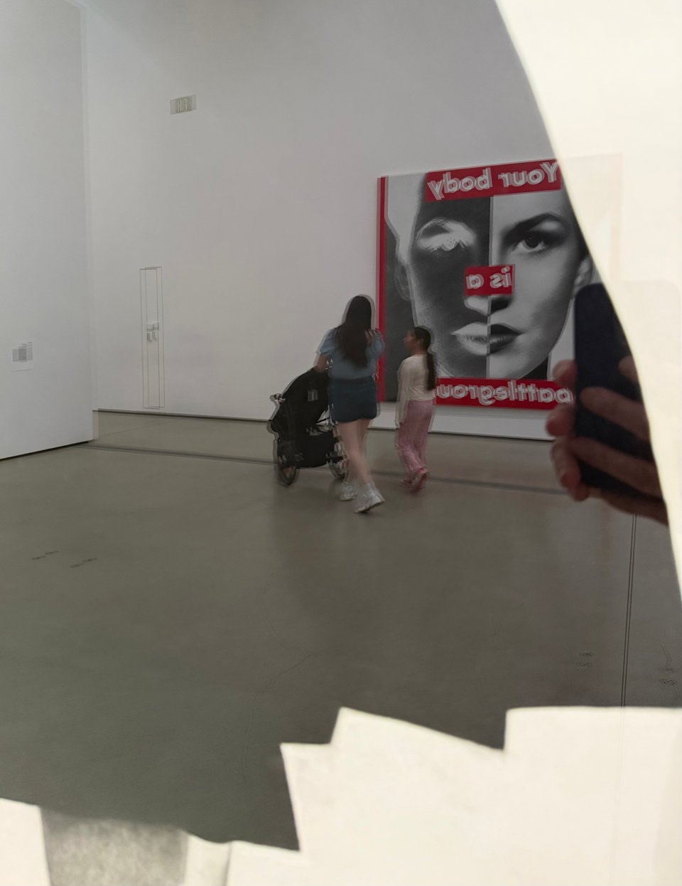Detail photo of Robert Longo's "Untitled (Men in the Cities: Ellen)," but the dark field of the skirt and the glass frame reflect a piece of art from the other side of the room, Barbara Kruger's "Your body is a battleground," as well as a mother pushing a pram while her older child walks beside her.