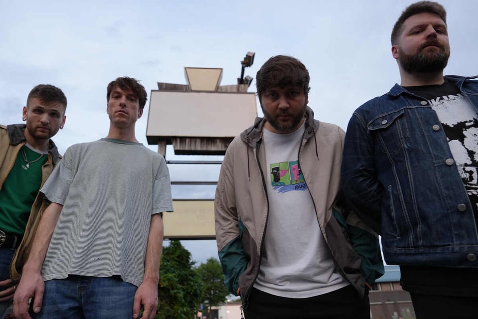 Four men stand in an uneven line in front of a vintage-style shopping center sign that is blank, looking into the camera.