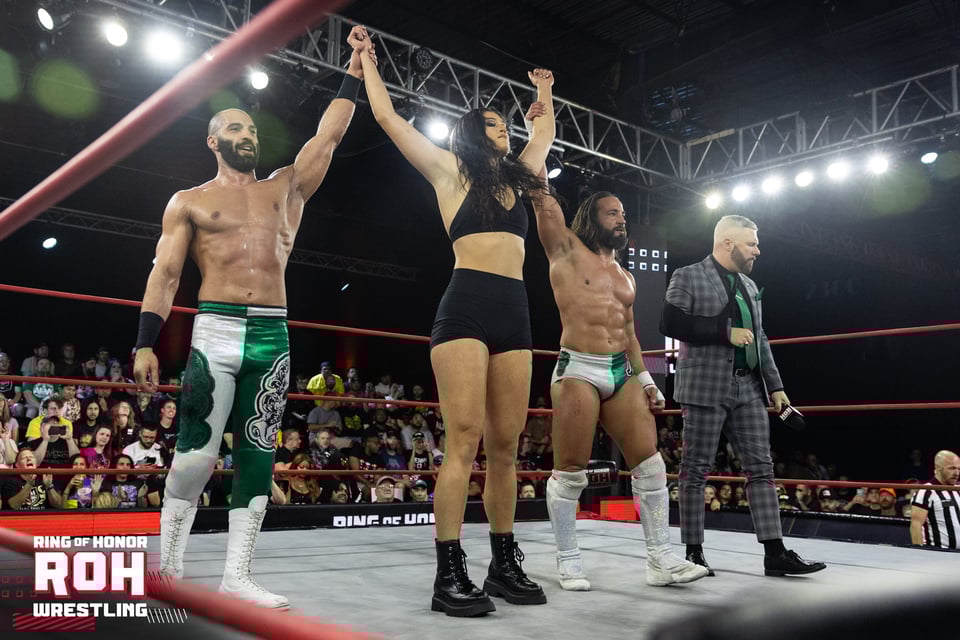 Stori Denali has her arms hoisted up triumphantly by two wrestlers in green-and-white costumes. It's presumed this is her presentation to her new audience.