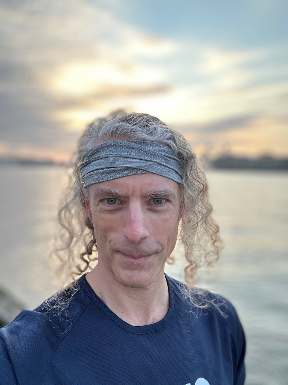 portrait mode selfie of white man in front of river in Rotterdam; he's wearing a blue tech-running shirt, and a light-gray headband, and has long curly hair and a smirk that doesn't betray how pissed of he is at his clients for texting at 2am and 4am while he was trying in vain to sleep before his big presentation later in the day